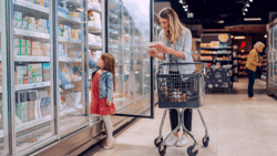 Photo of a woman and a young girl grocery shopping in the frozen foods aisle of a supermarket. The girl reaches into an open freezer door while the woman stands nearby holding a pack of sausages and examining the label, with a shopping cart in front of her. The setting is brightly lit, with other shoppers visible in the background.